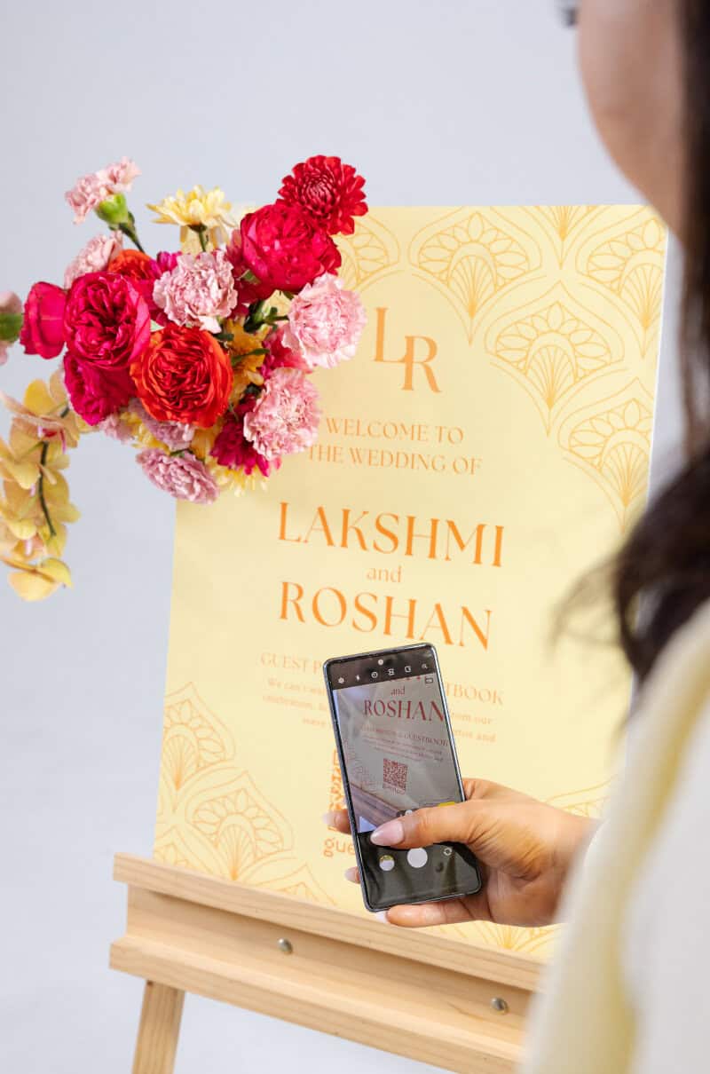Une femme prend une photo avec son smartphone d'un panneau de bienvenue orné d'une composition florale de fleurs roses et rouges, lors d'un mariage indien.