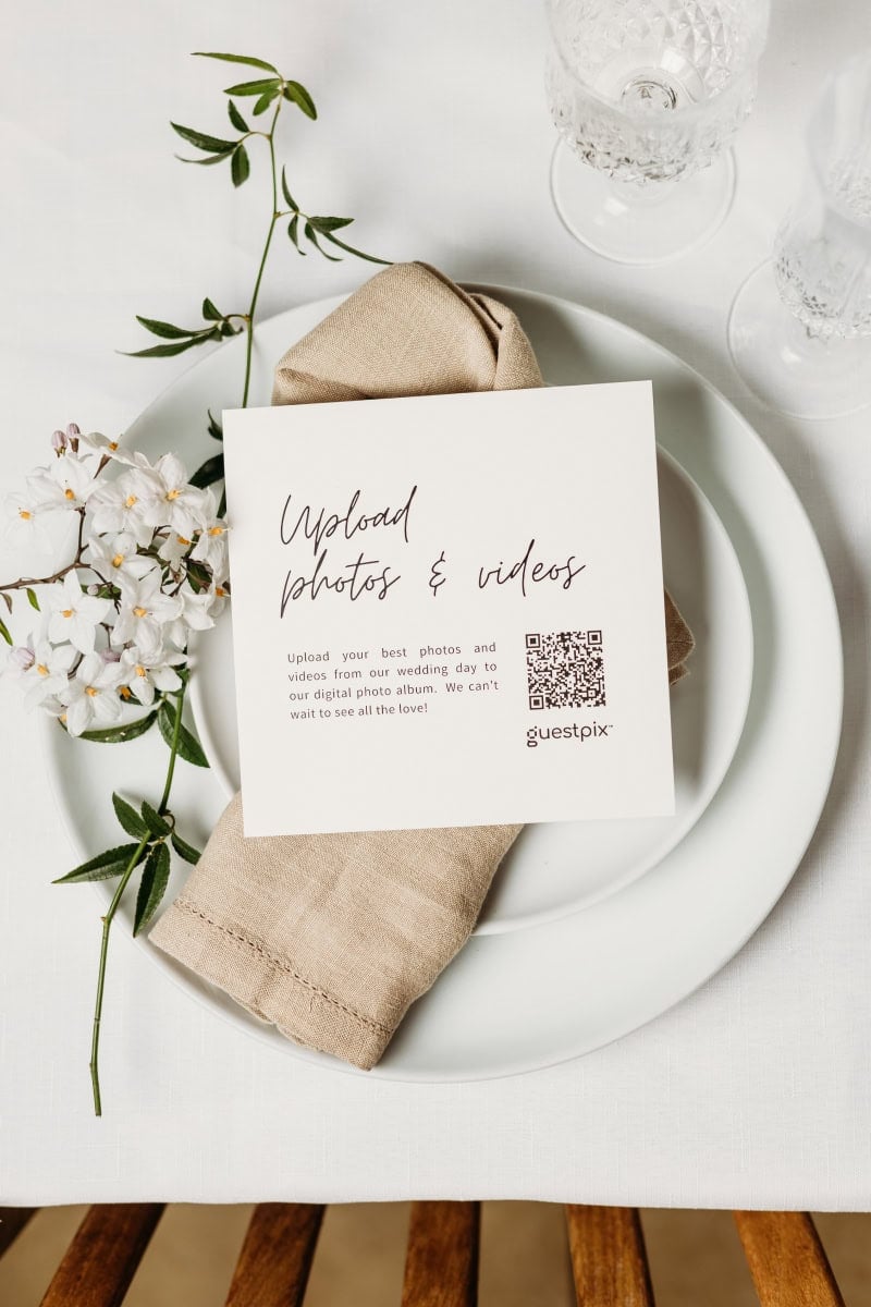 Une élégante composition de mariage composée d'une carte blanche ornée d'un texte imitant l'écriture manuscrite, d'un code QR et d'une serviette beige, le tout disposé sur une assiette blanche rehaussée de touches florales, pour un style moderne et minimaliste.