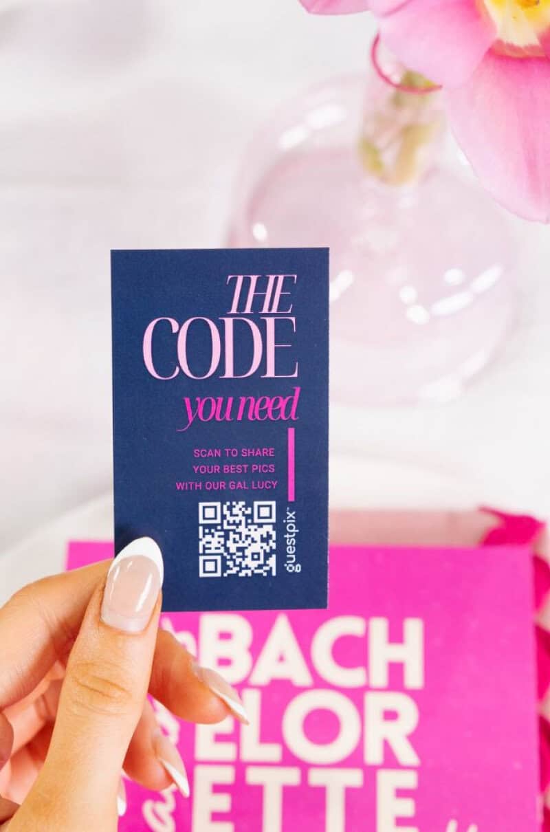 Primo piano di una mano che regge un cartoncino con un codice QR per la condivisione delle foto dell'evento alla Bachelorette Suite, con fiori rosa e un cartello rosa sullo sfondo.