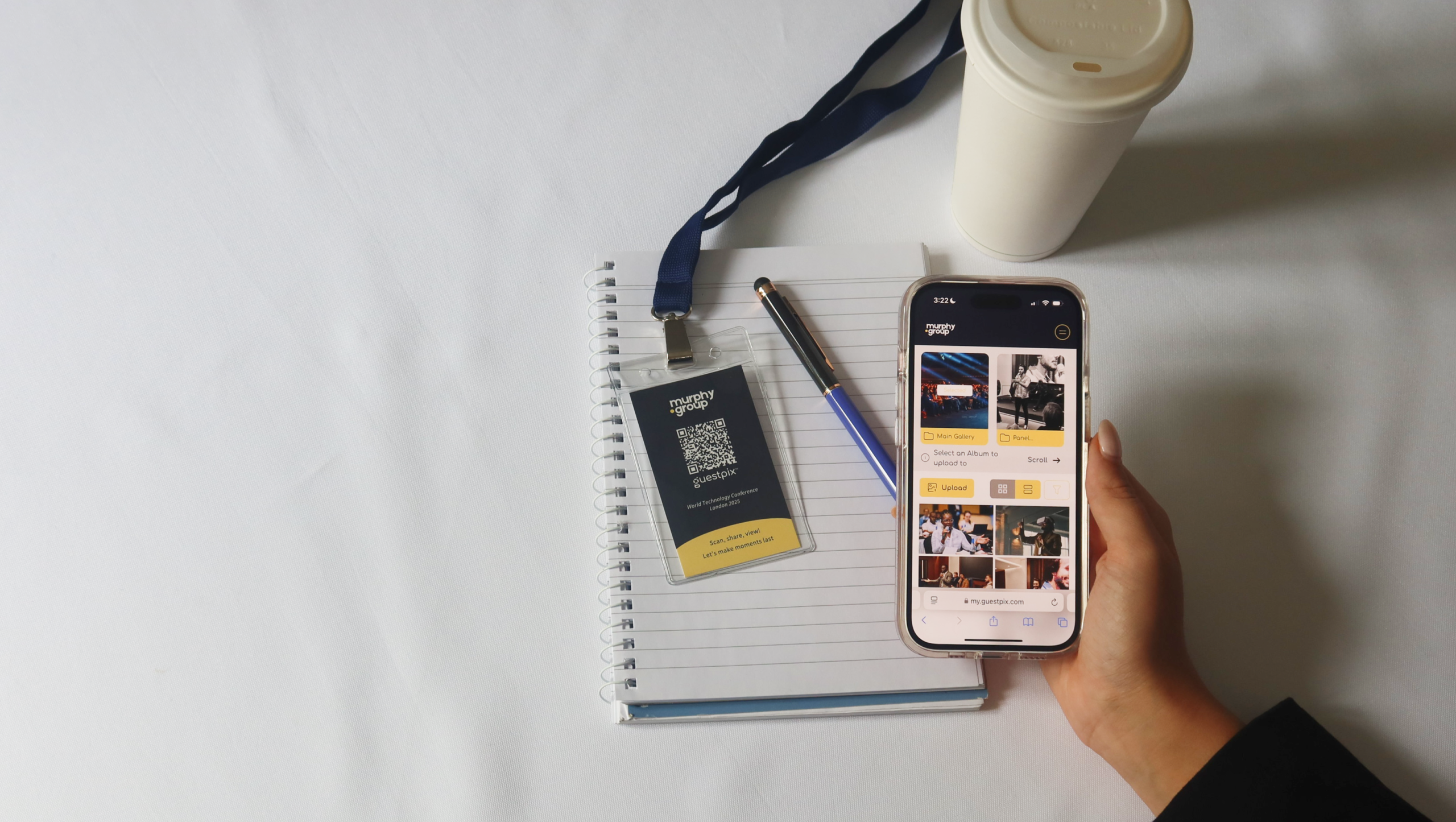 Eine Hand hält ein Smartphone, auf dem eine Fotogalerie zu sehen ist, neben einem Spiralblock, einem blauen Ausweis mit Umhängeband, einem Stift und einer Tasse auf einer weißen Tischplatte.