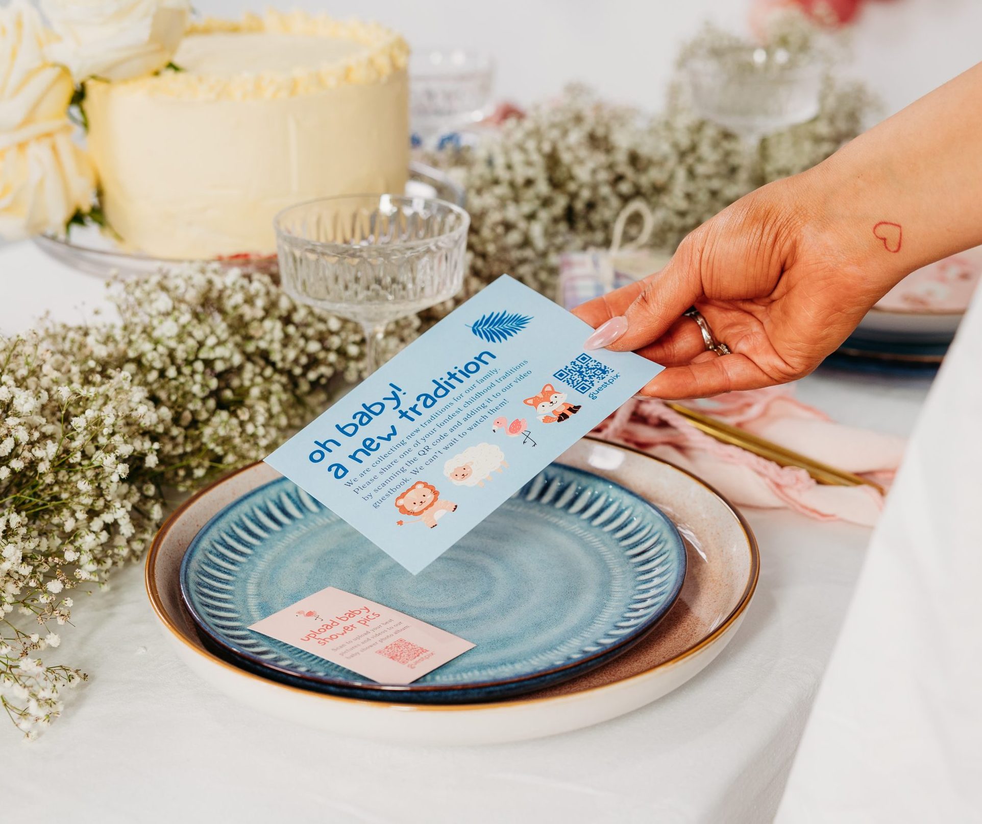 Primer plano de una mesa de boda con una tarta de color crema, flores blancas y una mano que sostiene una tarjeta azul claro sobre un plato con el borde azul.