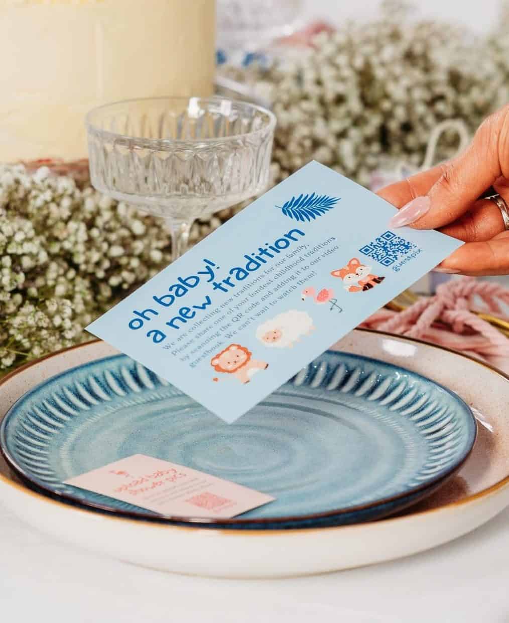 Tarta de boda con glaseado de nata y rosas blancas sobre una mesa con decoración floral y una mano que sostiene una tarjeta de invitación azul.