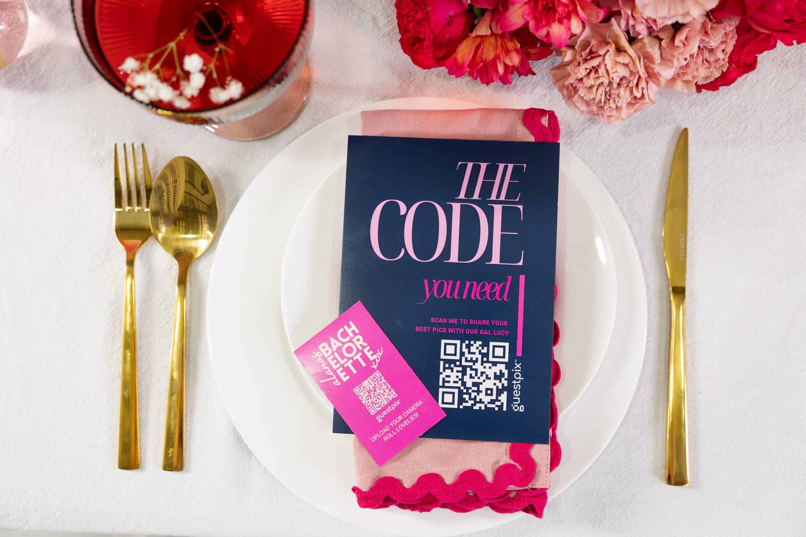 Tavola apparecchiata con un piatto bianco, posate dorate, un tovagliolo rosa e un opuscolo blu scuro con la scritta “THE CODE you need”, un codice QR e dei fiori rosa in alto.