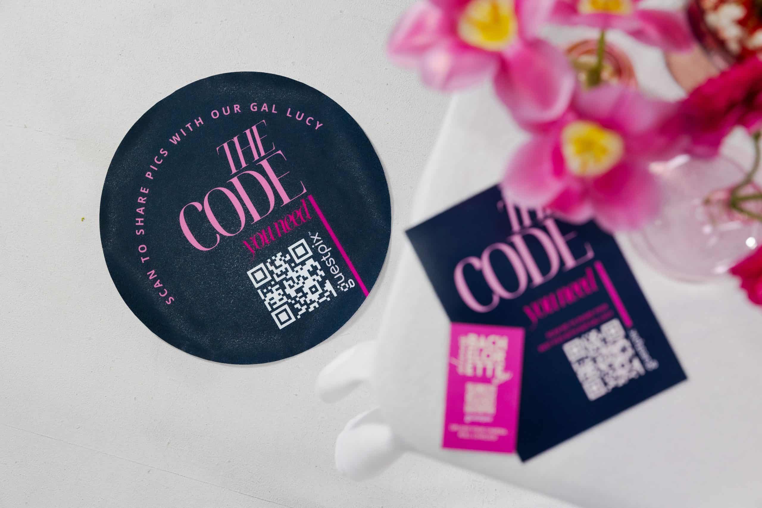 Sous-verre rond noir avec l'inscription « THE CODE » en rose et un code QR, à côté de fleurs roses dans un sac blanc.