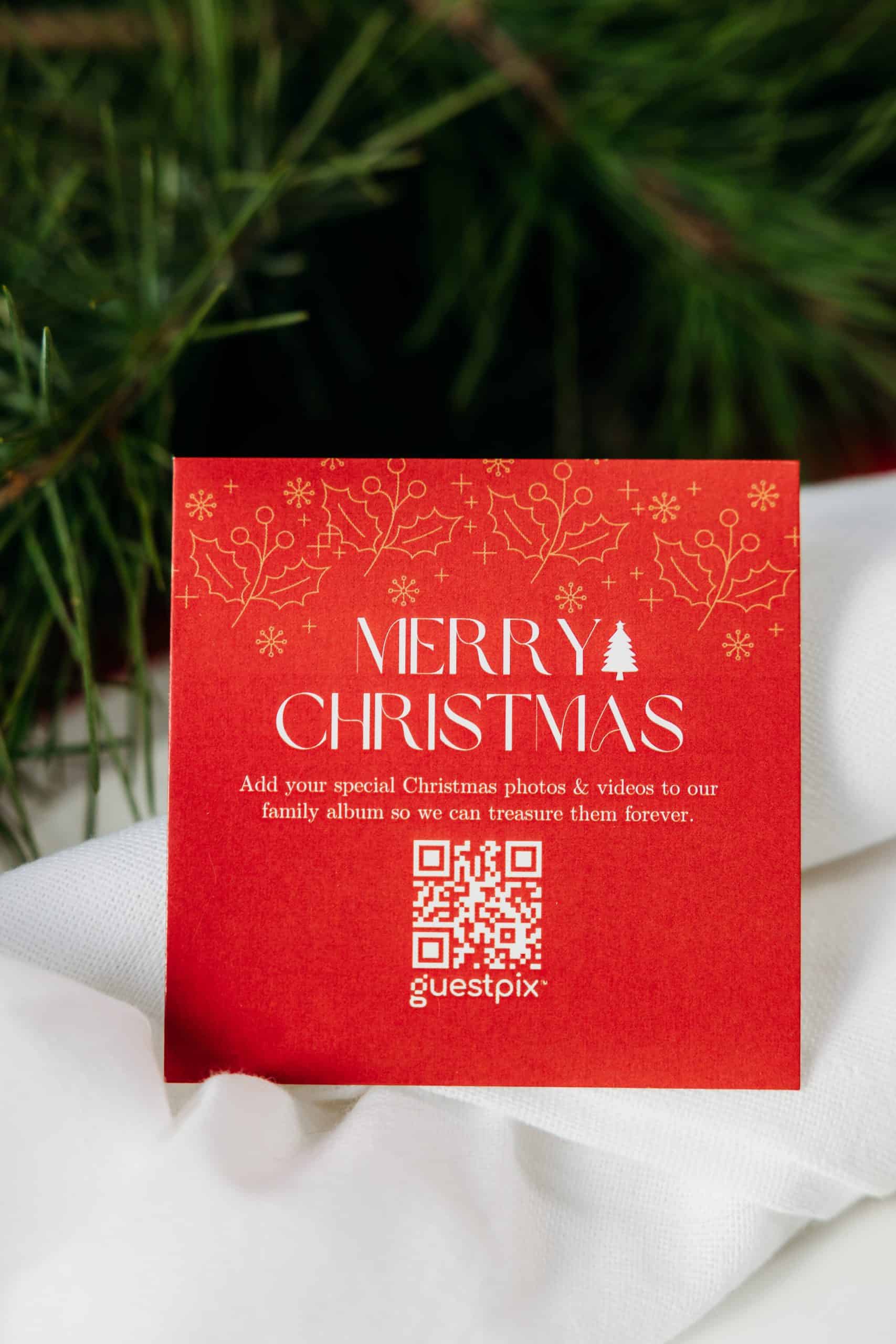 ヒイラギの装飾と白い文字で「MERRY CHRISTMAS」と書かれた赤いクリスマスカード。カードにはQRコードが印刷されており、背景には緑の葉が散りばめられた白い布が写っている。