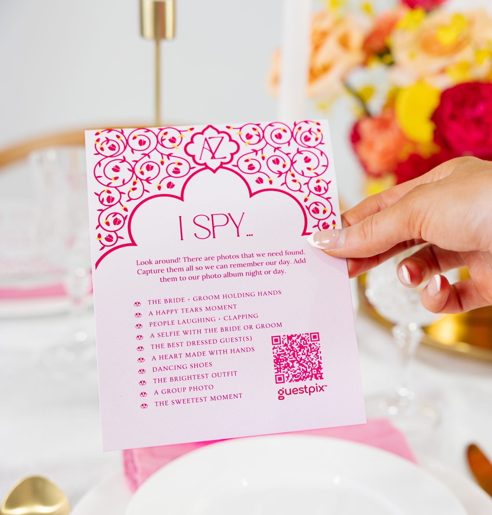 Tarjeta de invitación rosa de «I SPY» sostenida por una mano en una mesa festiva con platos blancos y flores de color amarillo brillante en un fondo difuminado.