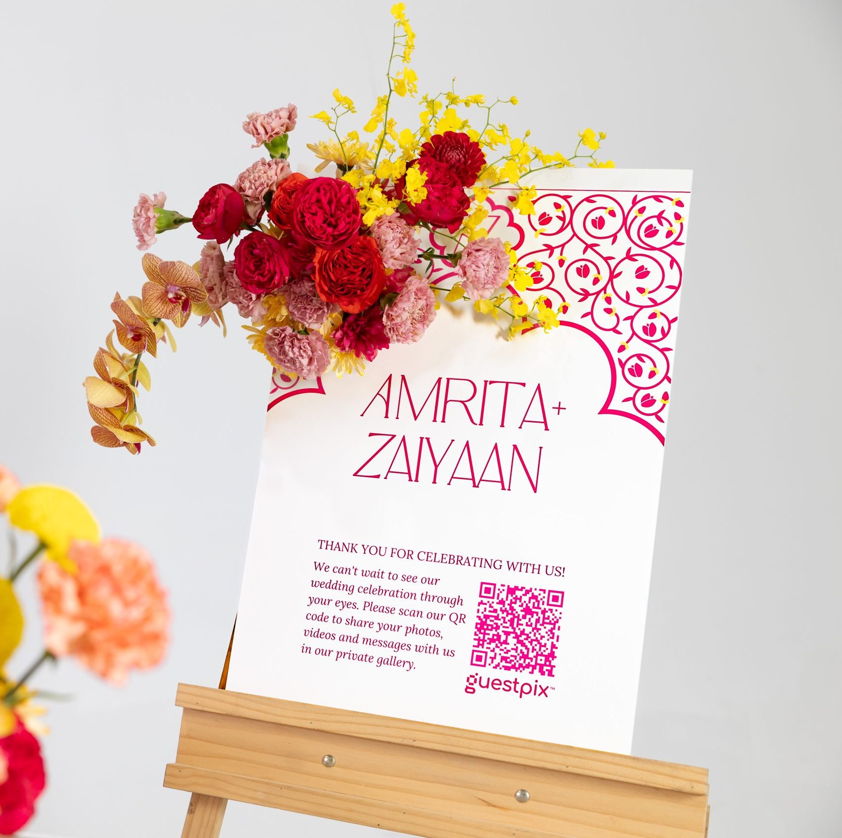 Woman in a pink dress holds a smartphone beside vibrant flower arrangements and a white decorative card on a wooden stand.