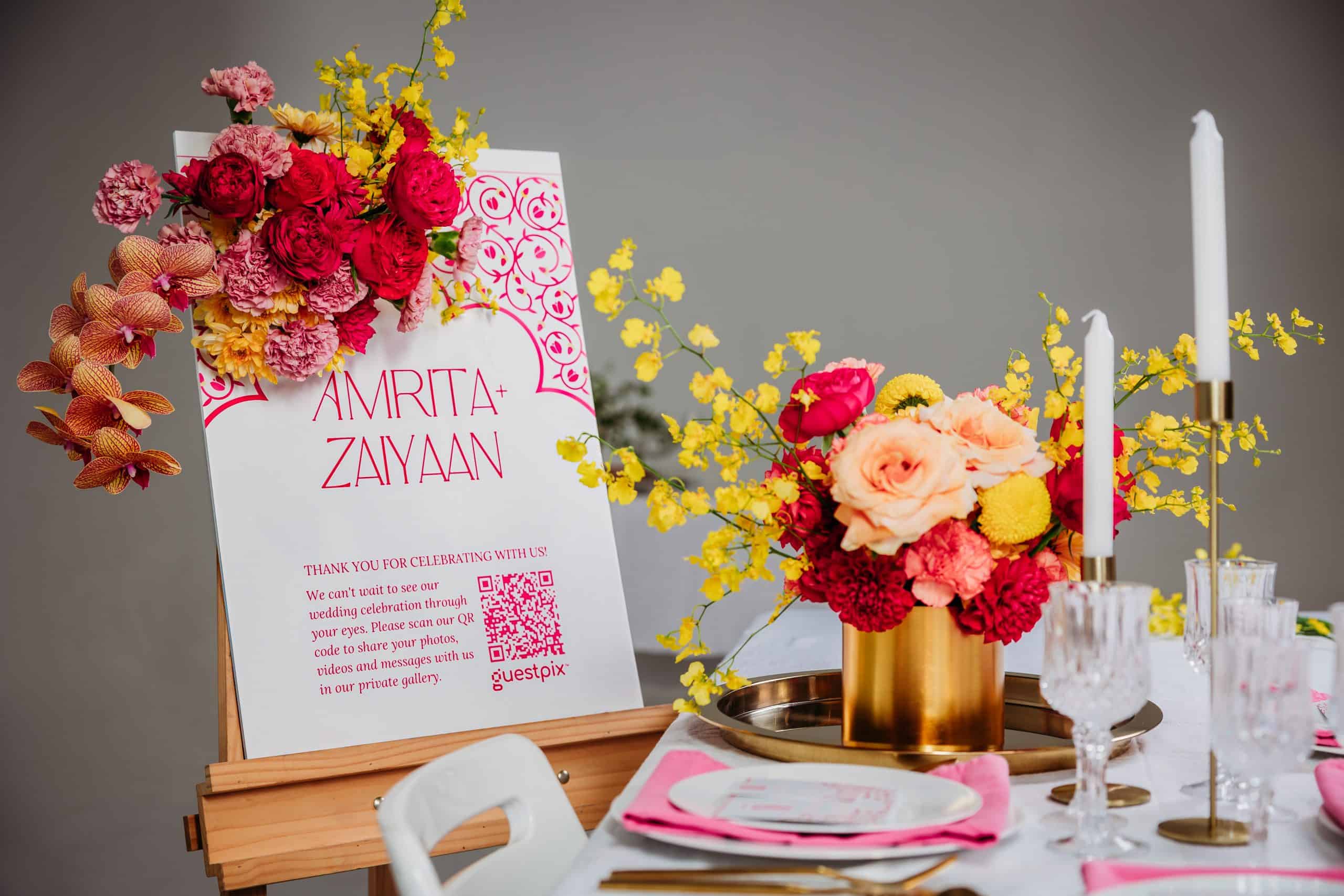 Cartel de bienvenida a una boda con flores de color rosa vivo y amarillo y un borde floral, en el que se lee «Amrita + Zayan», sobre una elegante mesa decorada.