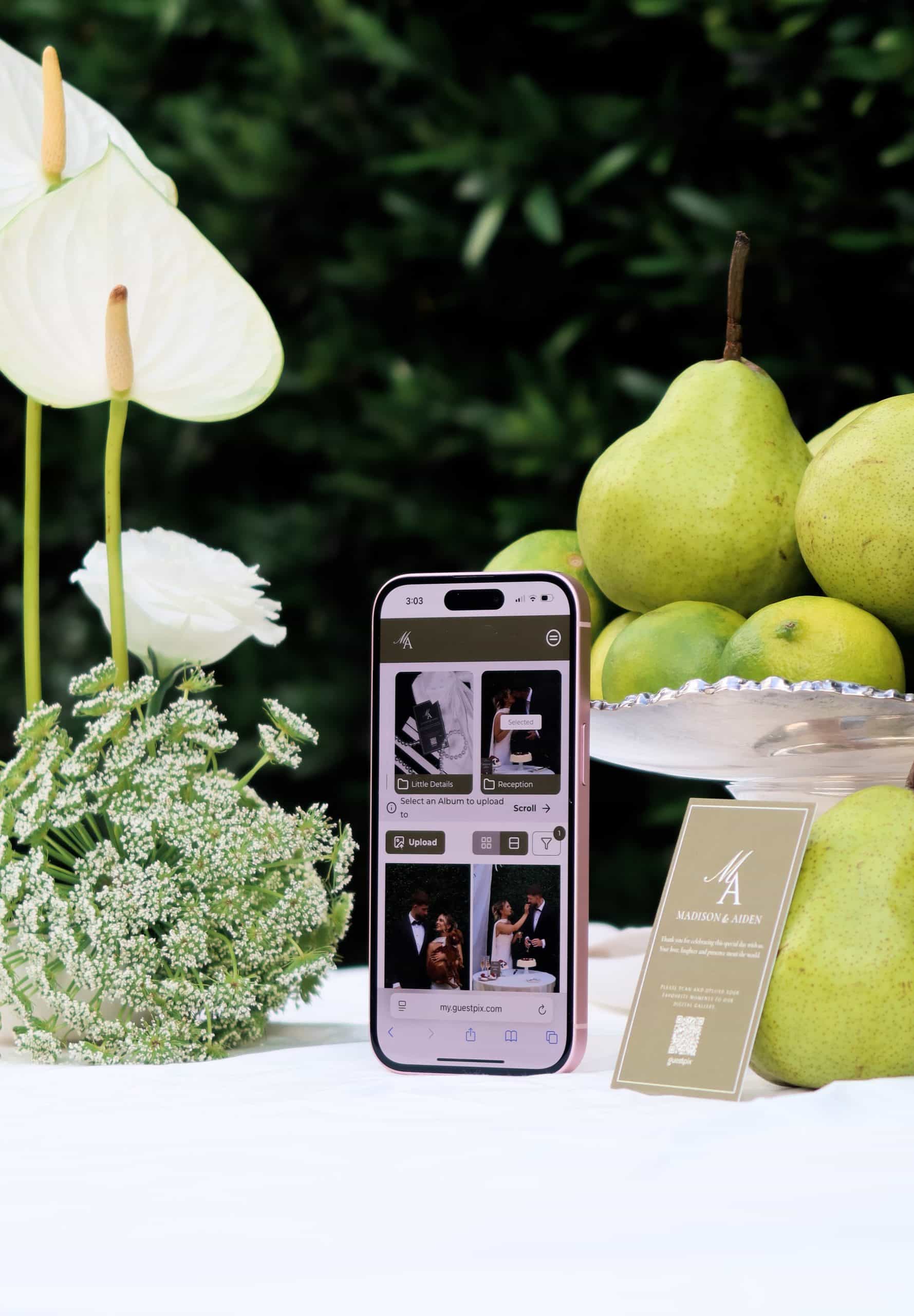 Wedding QR code sharing at an elegant event, featuring a smartphone displaying the GuestPix app with wedding photos, surrounded by fresh flowers and pears for a stylish wedding photo setup. - GUESTPIX | #1 QR Code Event Photo Sharing Platform Close-up of a smartphone displaying the GuestPix app on a wedding table, surrounded by elegant white flowers, green pears, and a wedding invitation, illustrating digital photo sharing at a wedding event.