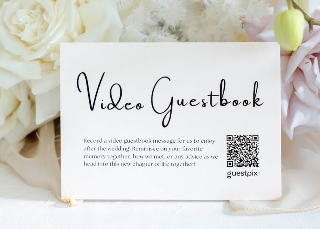 A wedding video guestbook card with a QR code, surrounded by soft white and blush flowers, inviting guests to record video messages and preserve wedding memories digitally.