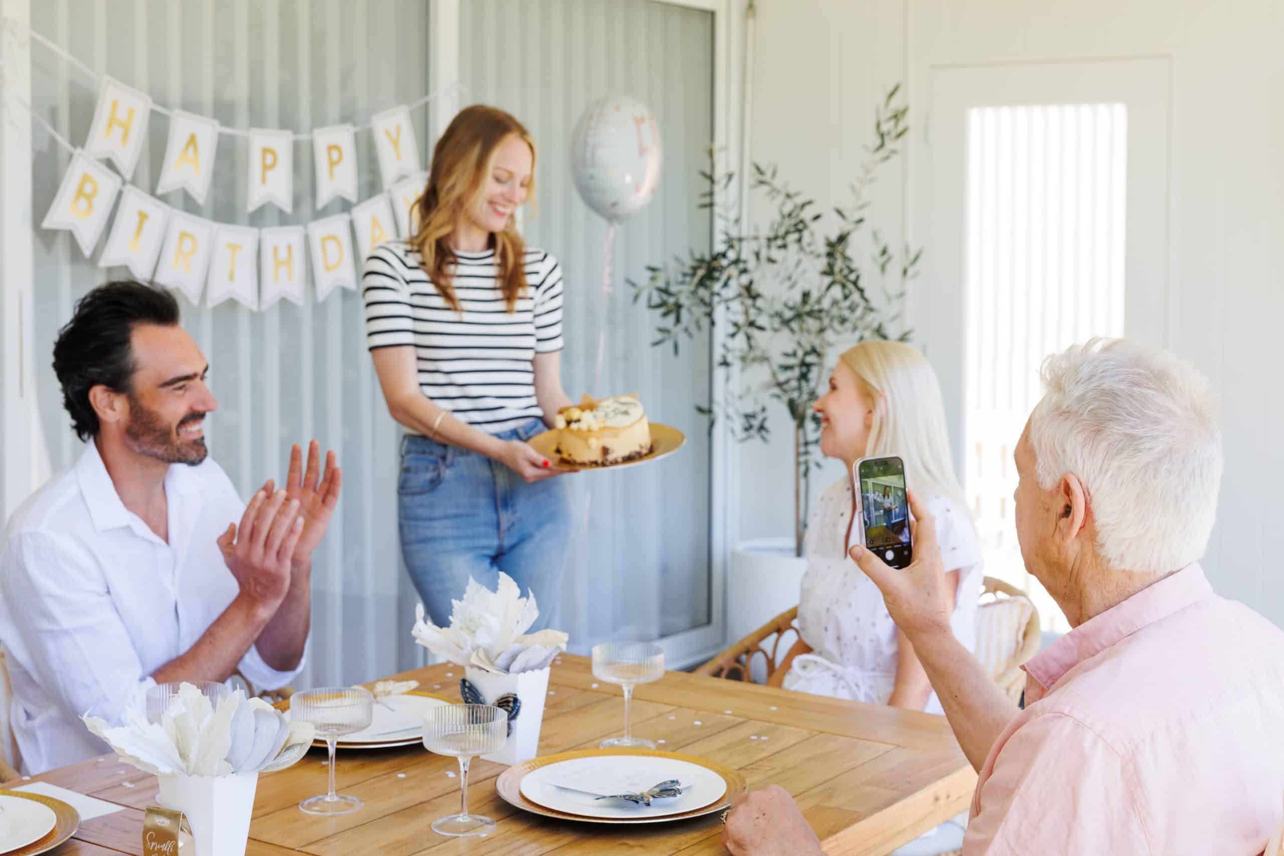 Fiesta de cumpleaños en la que una mujer presenta una tarta mientras los demás aplauden y sonríen en una habitación luminosa.