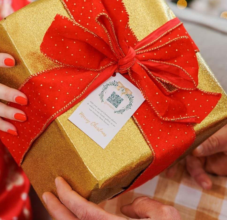 赤いクリスマス衣装を身にまとった女性が、赤いリボンが結ばれた金色のプレゼントを誰かに手渡している。背景には、お祝いの雰囲気漂うぼやけた明かりが映っている。