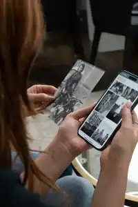 Person holds a black-and-white photo in one hand and looks at a smartphone screen showing a grid of grayscale image thumbnails.