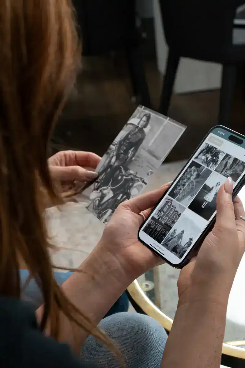Una persona tiene in una mano una foto in bianco e nero e guarda lo schermo di uno smartphone su cui è visualizzata una griglia di miniature di immagini in scala di grigi.
