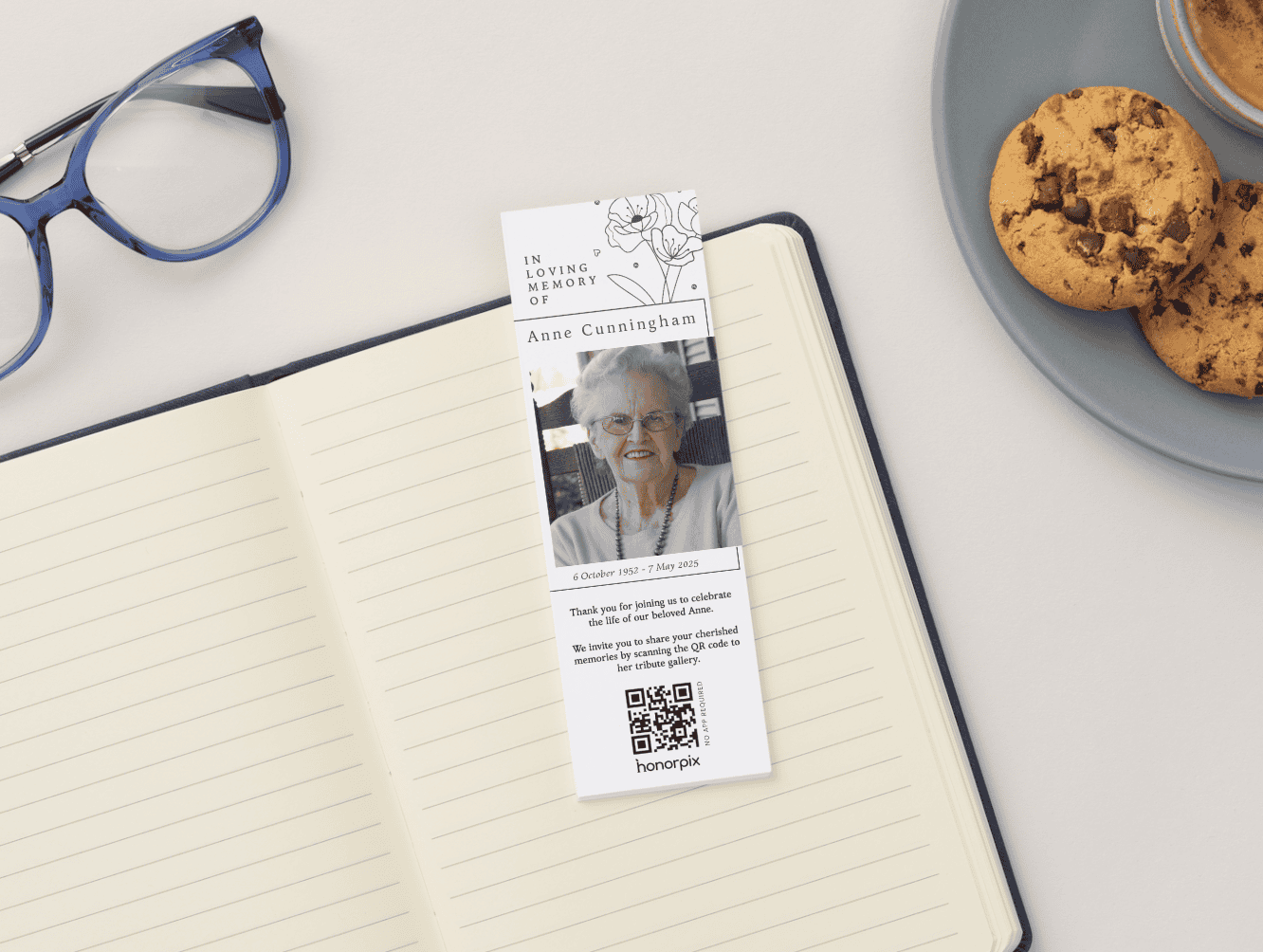 Marcapáginas conmemorativo con un retrato y un texto homenaje a Anne Cunningham, colocado sobre un cuaderno abierto junto a unas galletas y unas gafas azules.
