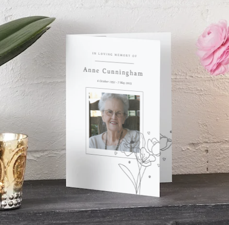 Carte commémorative représentant une photo posée sur une table en bois foncé, encadrée d'orchidées roses et d'un vase de fleurs roses, avec une lampe sphérique lumineuse à proximité.