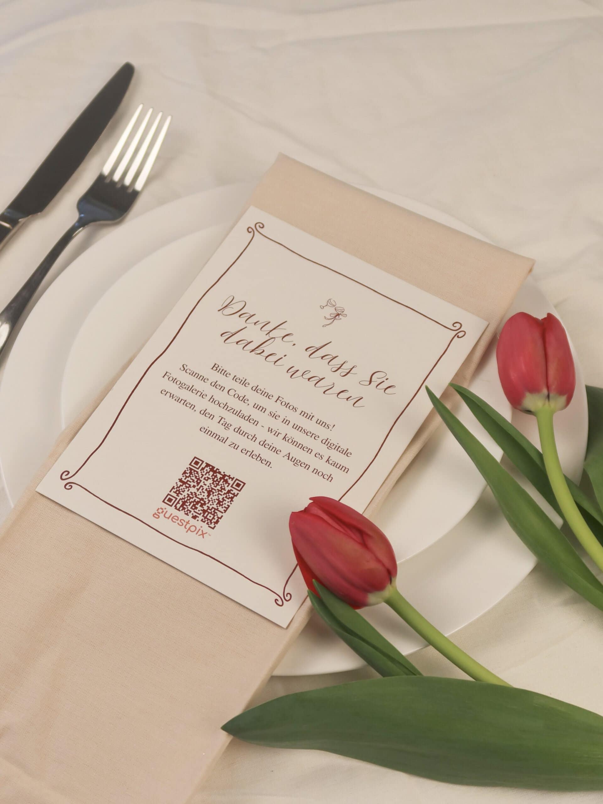 Tavola apparecchiata con coltello e forchetta su un piatto bianco, un tovagliolo beige e tulipani rossi accanto a un biglietto stampato con un bordo decorativo e un codice QR.