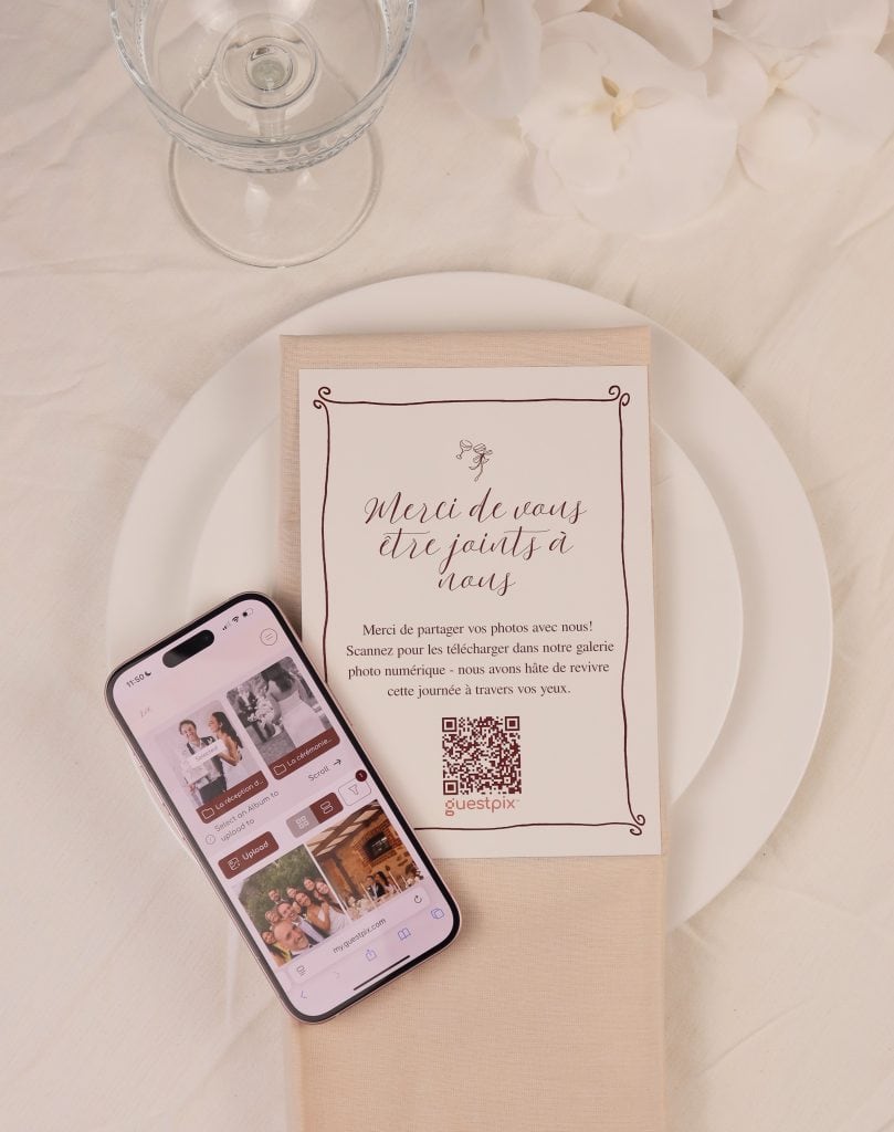 Tischdekoration für eine Hochzeit: weißer Teller mit beiger Serviette und einer bedruckten Karte mit einem QR-Code sowie ein Glas und ein Smartphone, auf dem Fotos auf einer hellen Tischdecke zu sehen sind.