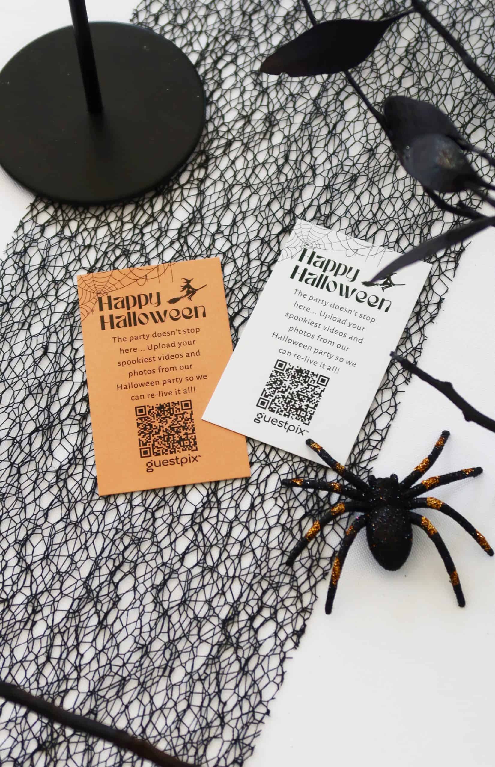 Due biglietti di Halloween (arancioni e bianchi) con la scritta "Happy Halloween", con codici QR su uno sfondo a ragnatela e una decorazione a forma di ragno nero nelle vicinanze.