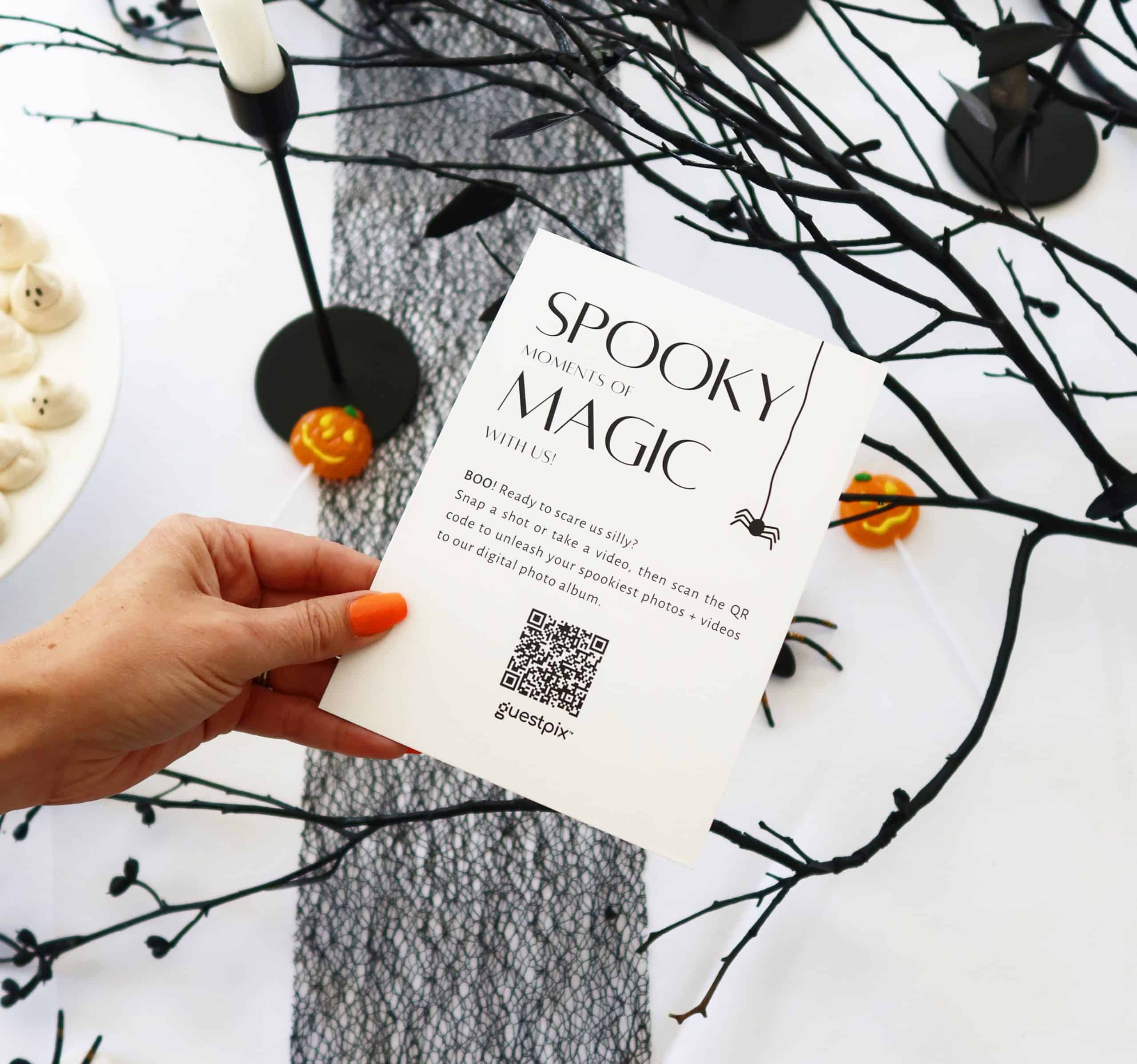 Mano che regge un volantino bianco di Halloween con un codice QR su una scrivania decorata con una ghirlanda di ramoscelli neri, piccole zucche e dolcetti arancioni.