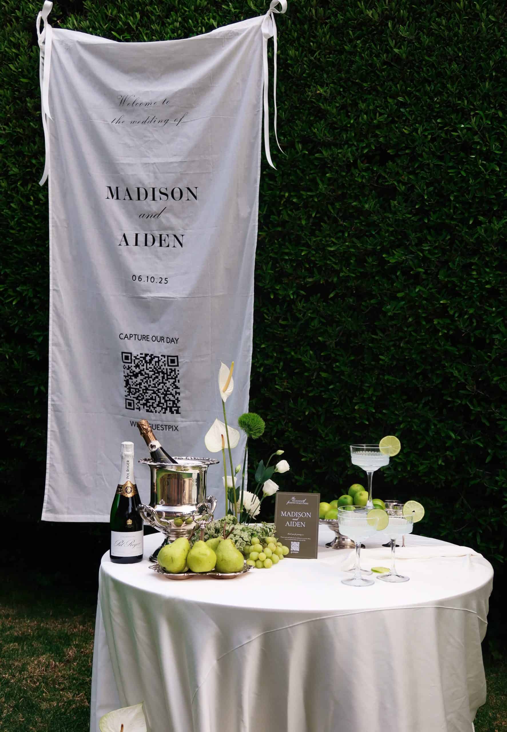 Wedding banner displaying couple's names Madison and Aiden with a QR code for guest photo sharing, alongside a table with champagne, green apples, and decorative elements at an outdoor wedding.