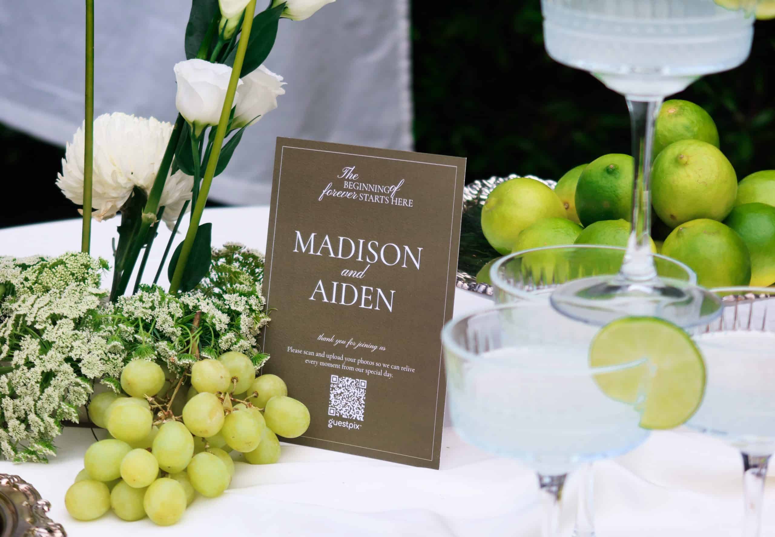 A stylish wedding table setup featuring green grapes, white flowers, and a QR code sign for guests to upload photos via GUESTPIX event platform.