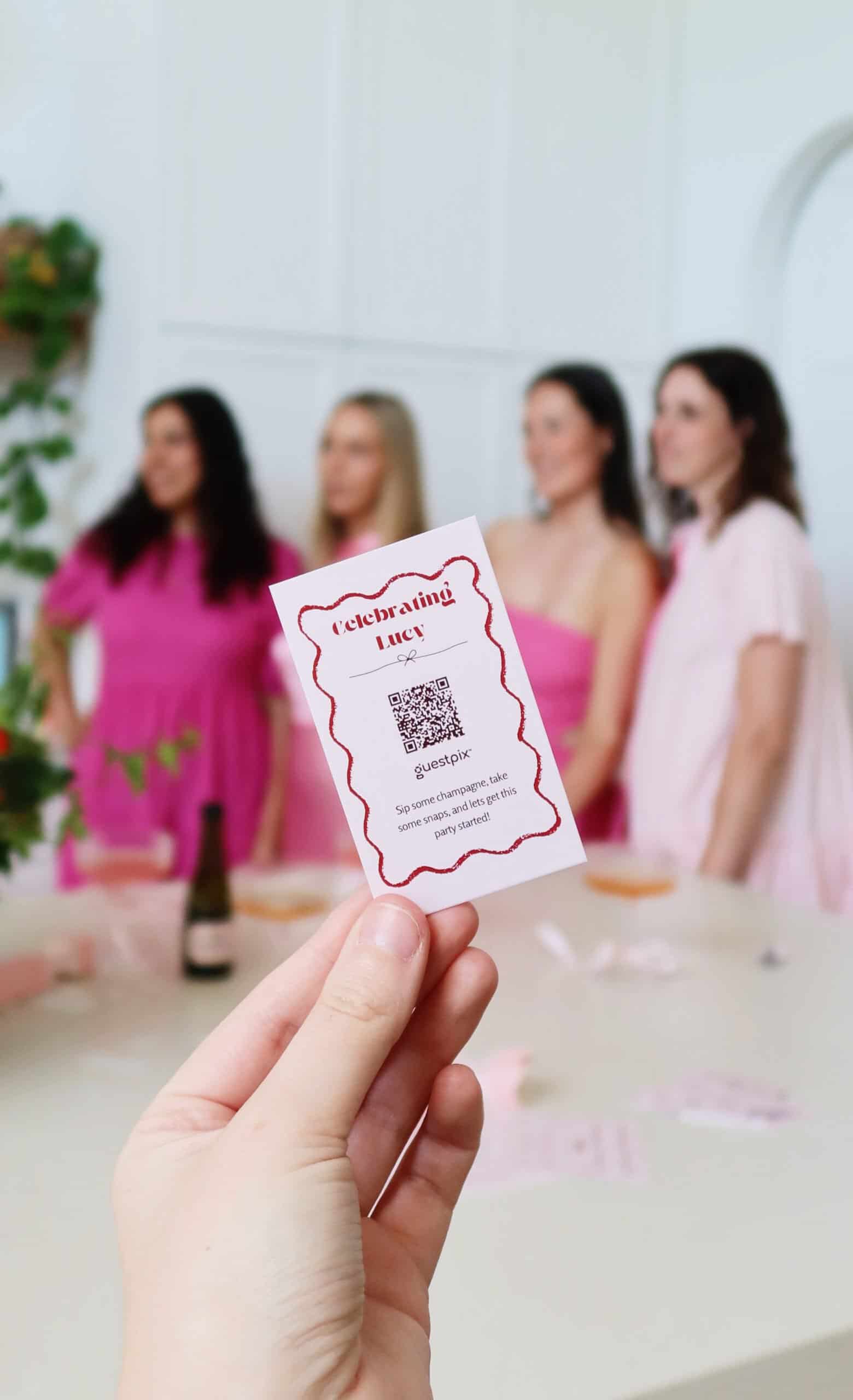 Una mano sostiene una entrada con un código QR para un evento festivo, con mujeres vestidas de rosa difuminadas al fondo. Esta imagen muestra la innovadora forma de compartir fotos de los invitados en las fiestas mediante Guestpix.