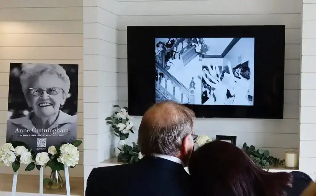 Memorial setup with a large Anne Cunningham portrait and white flowers as a couple watches a black-and-white video on a wall-mounted screen.