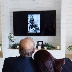 Zwei Personen sitzen mit dem Rücken zur Kamera und schauen auf einen Fernseher, auf dem ein Schwarz-Weiß-Foto einer Frau auf einem Motorrad vor einer Gedenkstätte mit Blumen und Kerzen zu sehen ist.