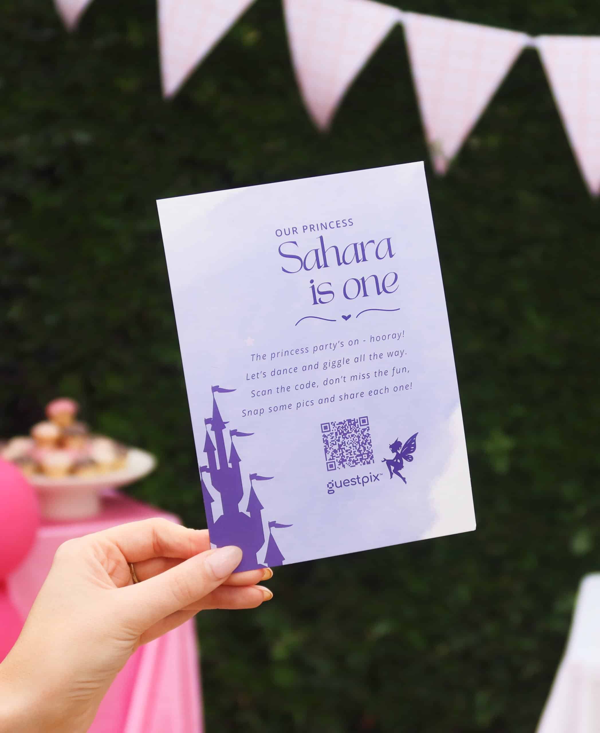 Una mano che regge un volantino per una festa a tema "principessa lavanda" con la scritta "Sahara compie un anno" e un codice QR, su uno sfondo rosa.