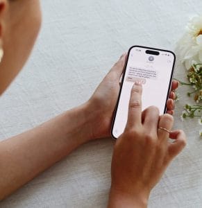 Nahaufnahme von Händen, die ein Smartphone halten, auf dessen Bildschirm eine SMS-Unterhaltung zu sehen ist; daneben stehen ein Leuchttisch und weiße Blumen.