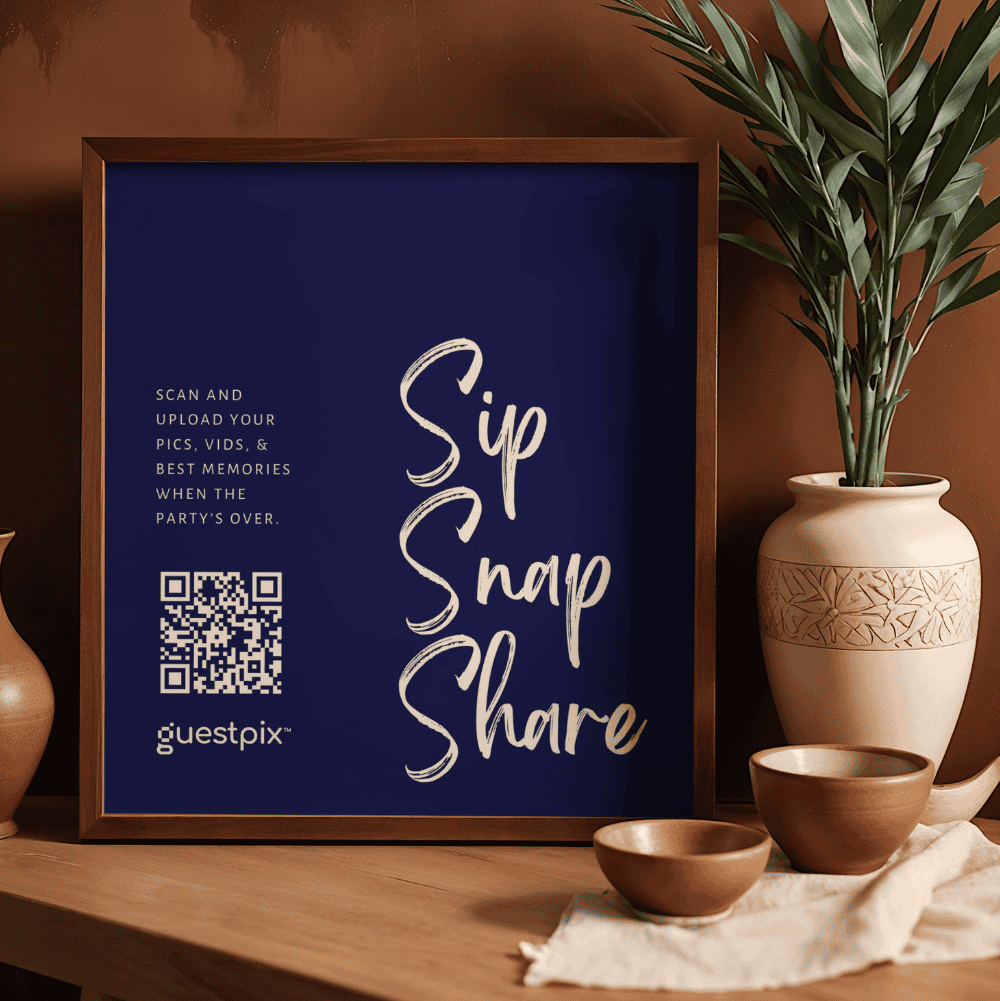 Cartel enmarcado de color azul oscuro con el texto «Sip Snap Share» escrito en letra caligráfica blanca, un código QR y el logotipo de «guestpix» a la izquierda, colocado en una estantería de madera junto a una pieza de cerámica y una planta.