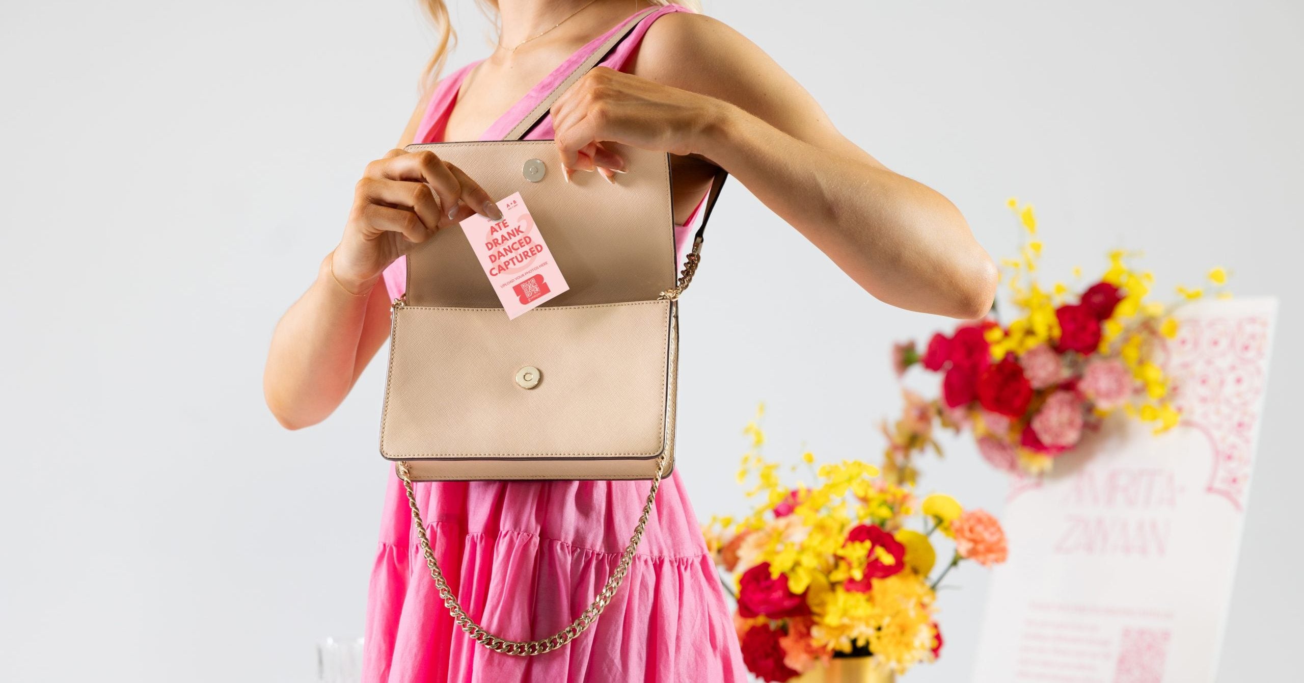 Mujer con un vestido rosa sosteniendo un bolso beige con una etiqueta de regalo rosa, sobre una mesa con flores al fondo.