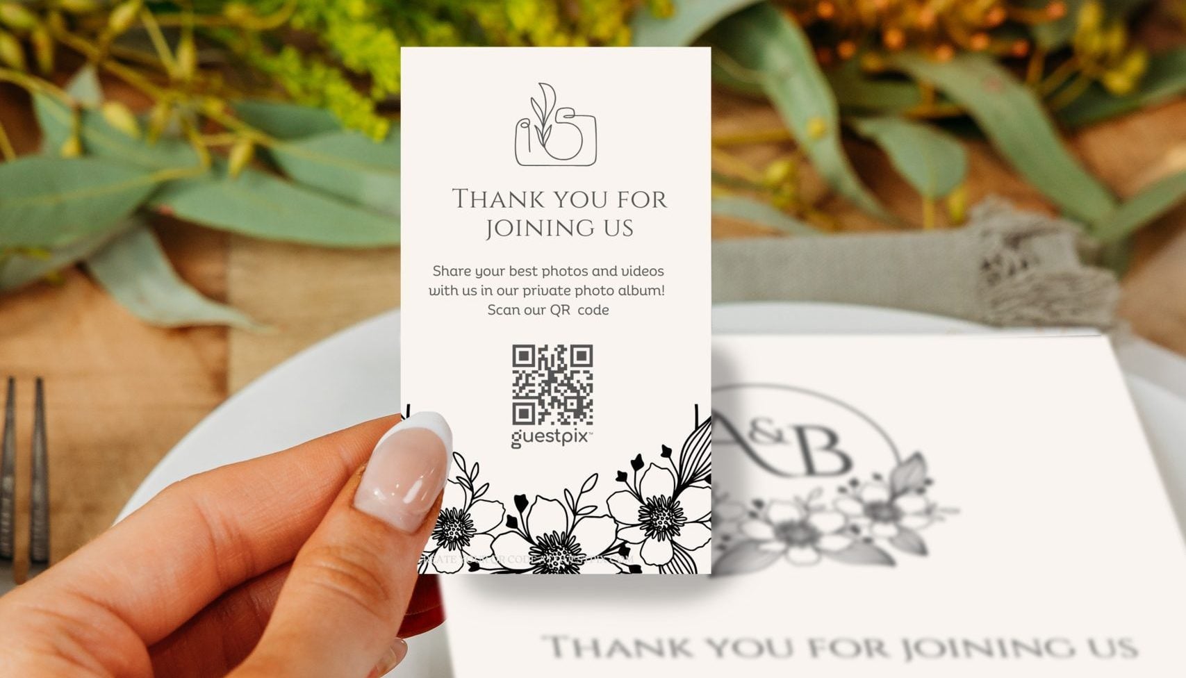Una persona que sostiene una tarjetita decorativa con un código QR sobre un plato llano en una mesa puesta; flores difuminadas al fondo.