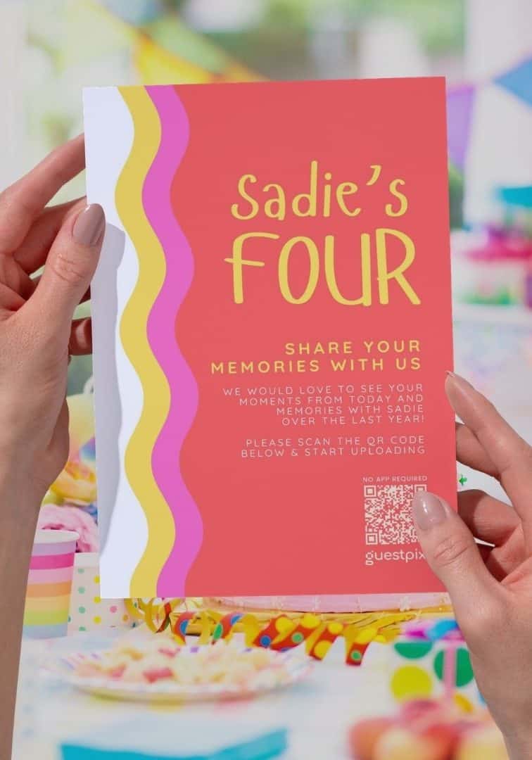 Primer plano de una colorida tarjeta de invitación a una fiesta en la que se lee «Sadie cumple cuatro», con un código QR, sostenida por dos manos, en una festiva celebración de cumpleaños con adornos y golosinas al fondo.