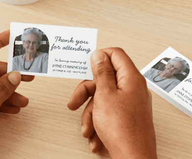 Des mains tiennent une carte de remerciement sur laquelle figure la photo d'une femme âgée et la mention « Merci de votre présence », posée sur une table en bois clair.