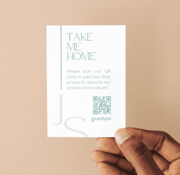 Weiße Karte mit der Aufschrift „TAKE ME HOME“ und einem QR-Code, die von einer Hand vor einem beigen Hintergrund gehalten wird (Guestpix-Branding).