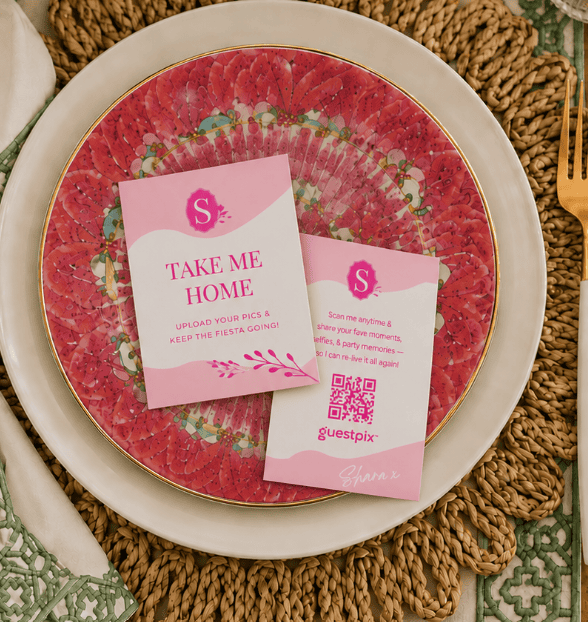 Des cartes d'invitation roses disposées sur une assiette décorative rouge et or, posée sur un set de table tissé ; l'une d'elles porte l'inscription « TAKE ME HOME », accompagnée du logo Guestpix et de motifs floraux.