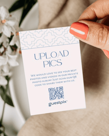 Tarjeta sujeta entre los dedos en la que se lee «SUBIR FOTOS», con instrucciones para subir fotos a través de un código QR para Guestpix; fondo floral suave.