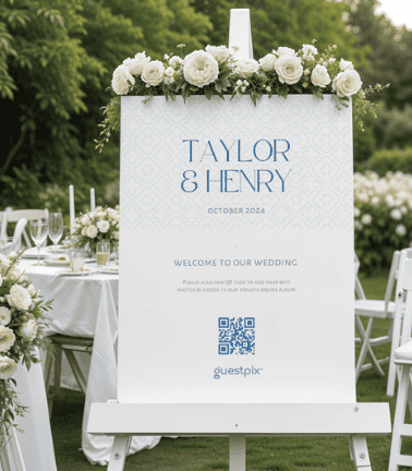 Cartel de bienvenida para una boda decorado con rosas blancas; en él se lee «Taylor &amp; Henry» y «Octubre de 2024», y lleva un código QR sobre un soporte.
