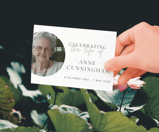 Primo piano di una mano che regge un biglietto commemorativo con la foto di una donna anziana e la scritta “In ricordo di Anne Cunningham”, con le date di nascita e di morte.