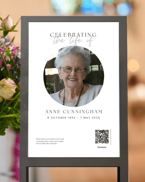 Poster commemorativo incorniciato su un supporto, con la foto di Anne Cunningham, il titolo “Celebrating the life of”, le date 1952–2025, un codice QR e dei fiori sullo sfondo.