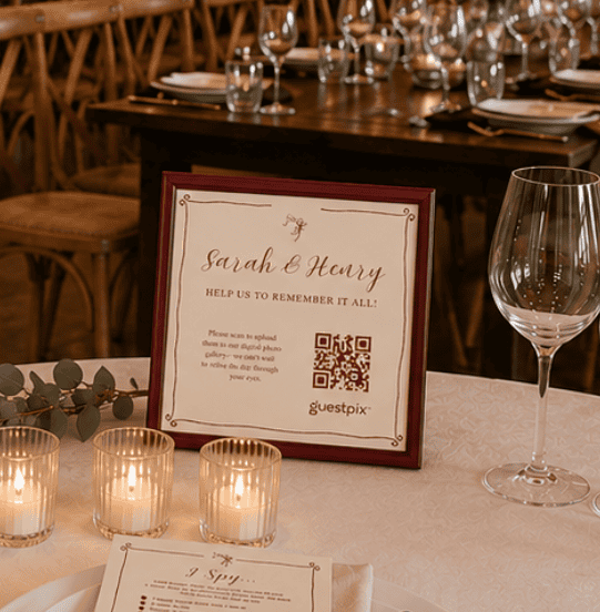 Panneau encadré portant les noms « Sarah &amp; Harry » et un code QR, posé sur une table de mariage, entouré de bougies votives et de verres.