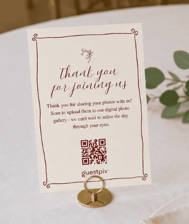 Carte de remerciement posée sur une table, ornée d'une bordure décorative, comportant un code QR permettant de télécharger des photos ainsi que le mot « guestpix », avec de la verdure en arrière-plan.
