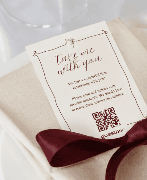 Carte d'invitation posée sur une serviette beige, nouée d'un ruban bordeaux, sur laquelle on peut lire « Emmène-moi avec toi » et qui comporte un code QR.