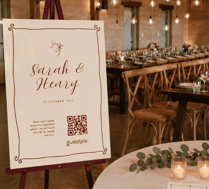 Panneau de mariage posé sur un chevalet, sur lequel on peut lire « Sarah &amp; Henry, 27 octobre 2026 », accompagné d'un code QR ; en arrière-plan, une salle de réception au style rustique avec des tables éclairées à la bougie.