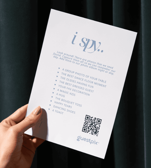 Eine Hand, die ein weißes „I Spy“-Spielblatt hält, auf dem eine Liste mit zu suchenden Gegenständen, ein QR-Code und das Guestpix-Logo zu sehen sind.