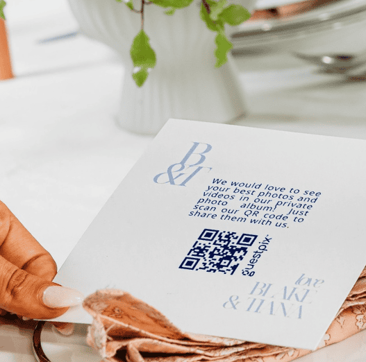 Primo piano di una mano che tiene in mano un biglietto d'invito bianco con un codice QR blu e un testo che invita a condividere foto e video.