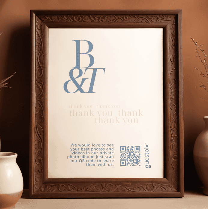Gerahmtes Schild mit großen blauen Initialen „B“ und „T“, dem wiederholt aufgedruckten Text „Danke“ und einem QR-Code für „Guestpix“ auf einem Holzrahmen.