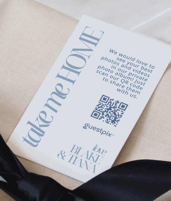 Weiße Einladungskarte mit dem Schriftzug „take me HOME“, einem QR-Code und dem Logo von Blake &amp; Tiana, die auf beigem Stoff liegt.